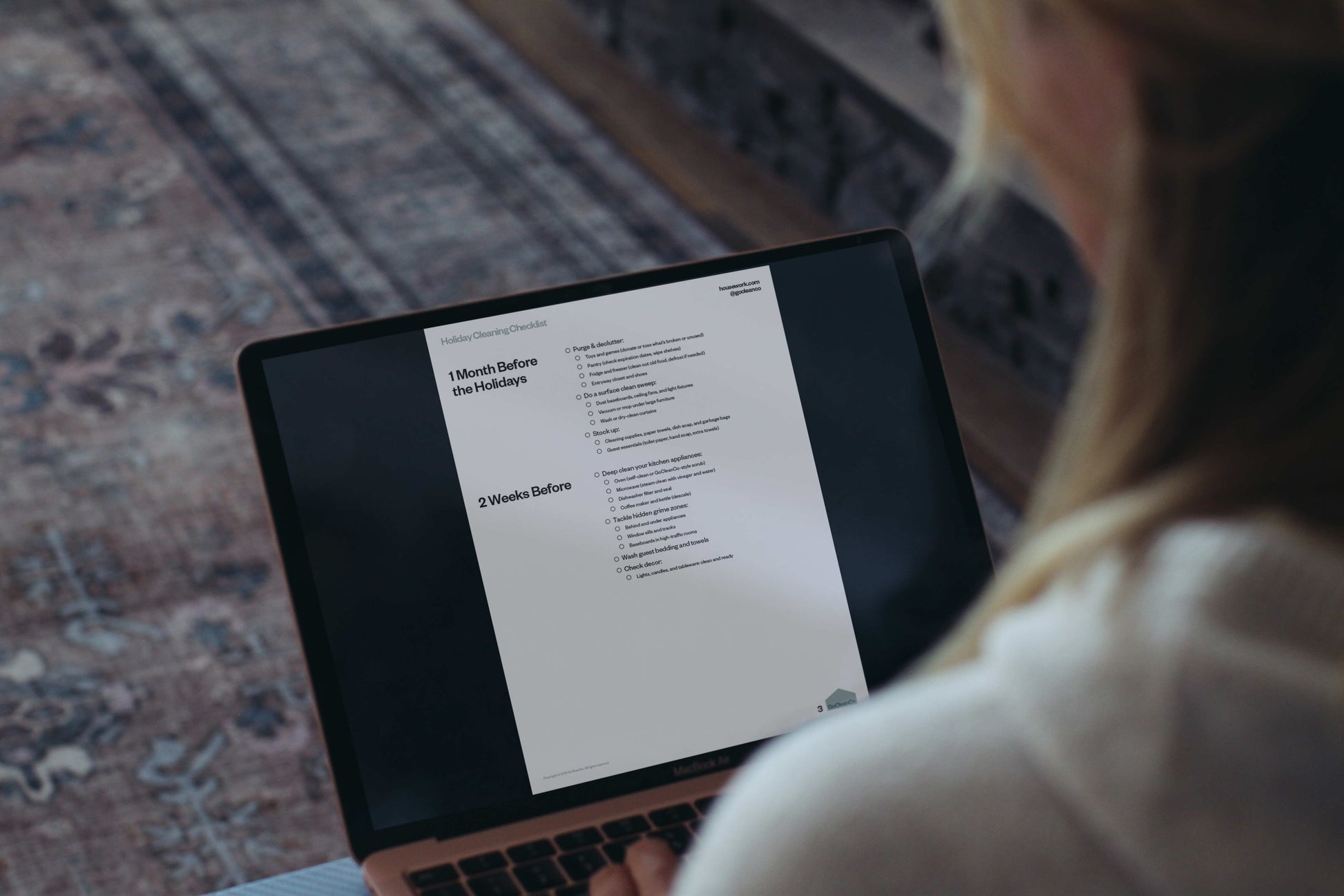 Person using a laptop with a Holiday Cleaning Checklist on the screen, sitting on a patterned rug.