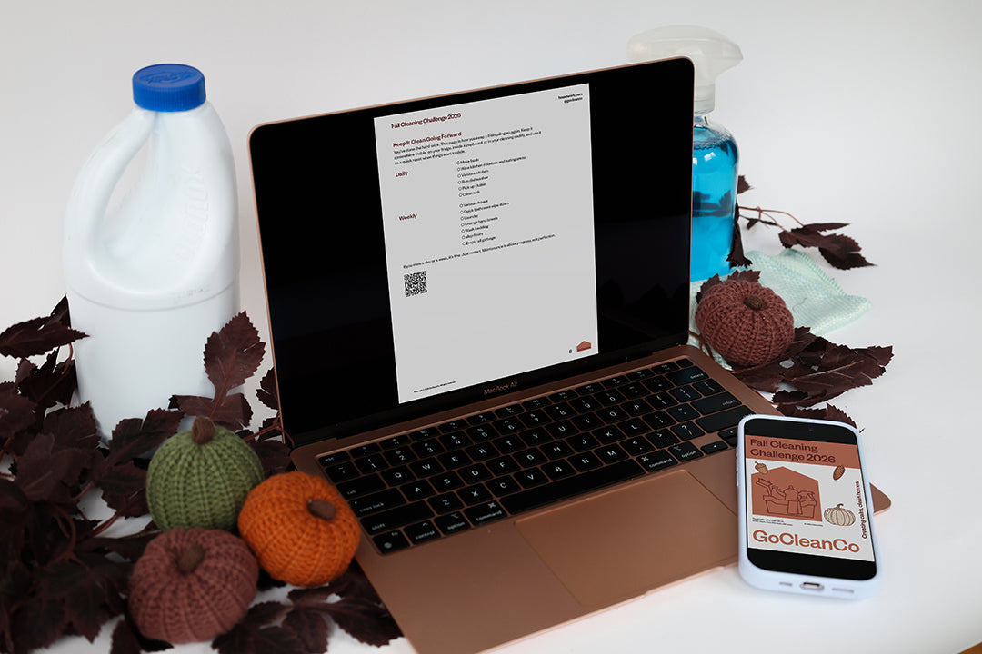 A laptop on a white surface displaying a cleaning challenge page, surrounded by fall-themed props including a bleach bottle, windex, knit pumpkins, leaves, and a smartphone showing the GoCleanGo Fall Cleaning Challenge.