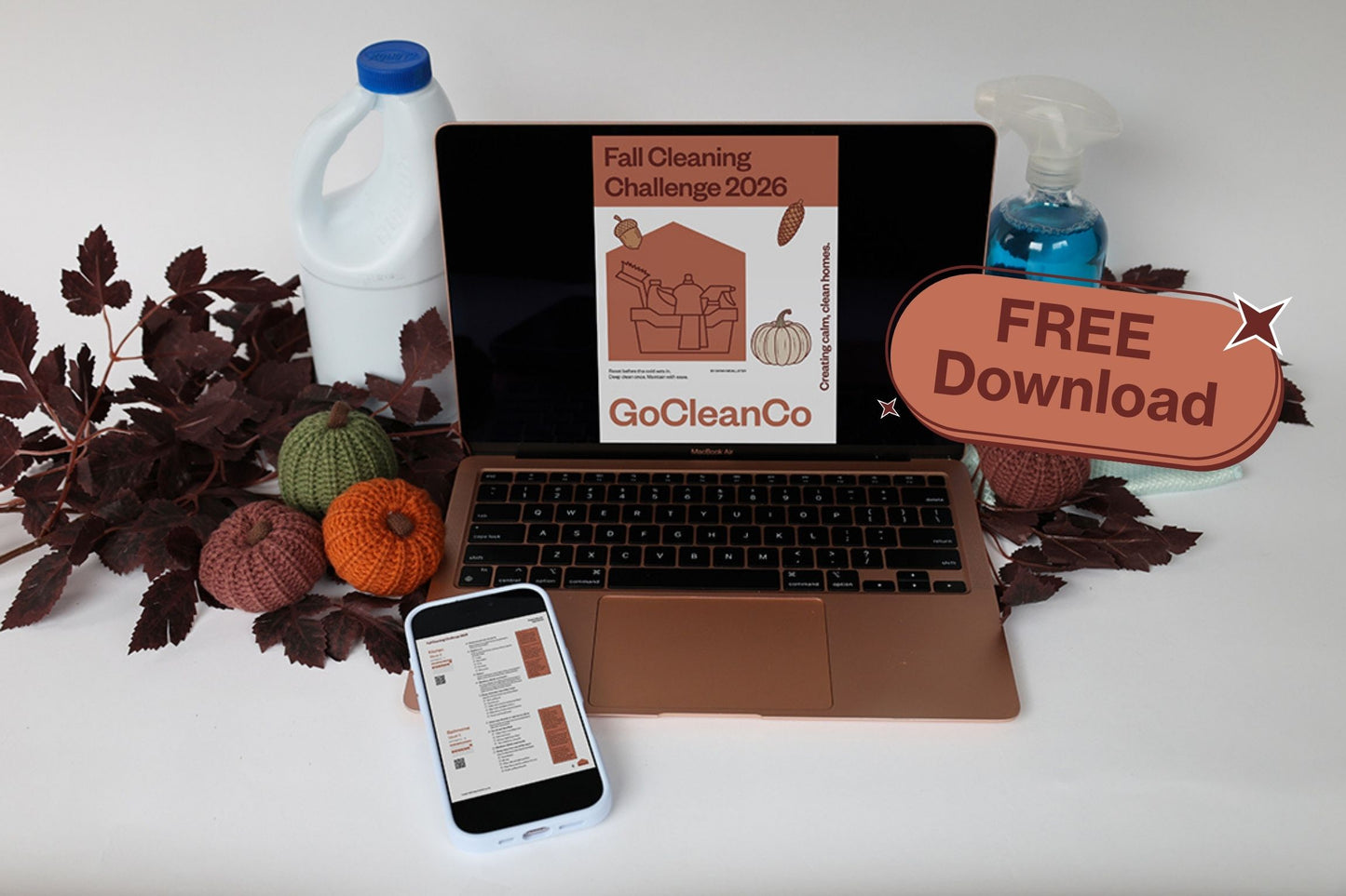 A laptop on a white surface displaying a fall cleaning challenge poster by GoCleanCo, surrounded by autumn-themed props including pumpkins, leaves, a bottle of laundry detergent, a spray bottle, and a phone. A brown badge on the right says FREE Download.