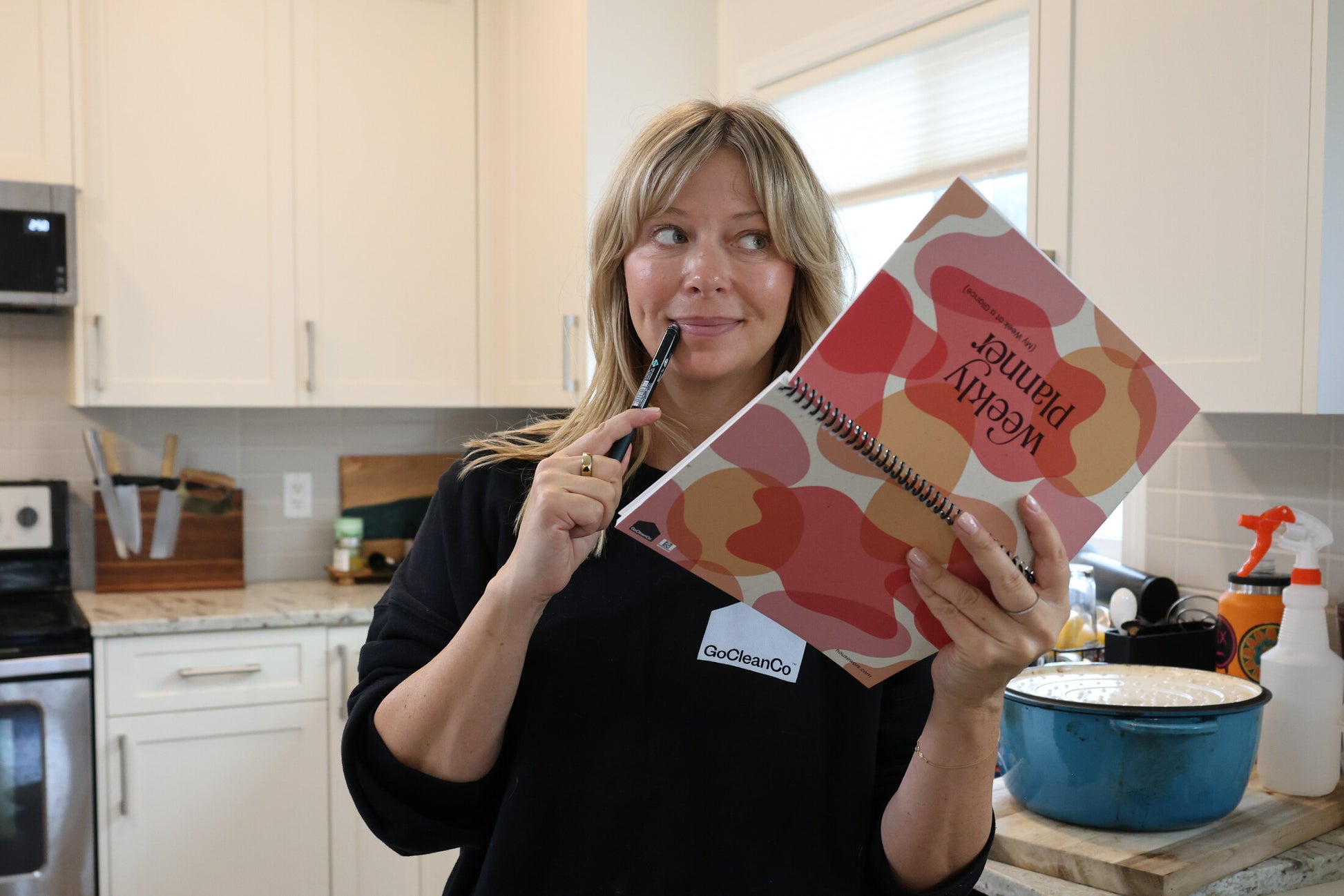 A woman in a kitchen holds a spiral-bound notebook titled 'Weekly Planner' with a colorful abstract pattern. She is wearing a black long-sleeve shirt with a white 'GoCleanCo' logo on the chest.