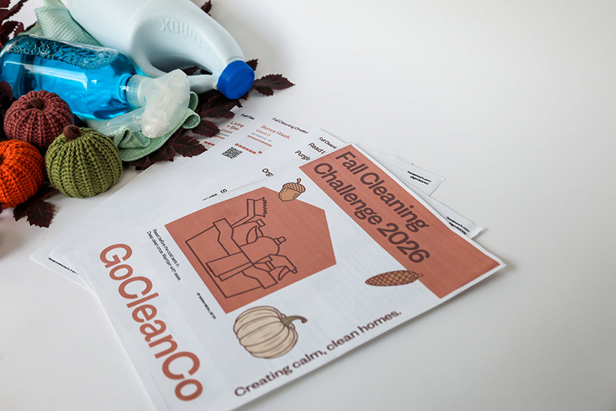 A GoCleanCo Fall Cleaning Challenge 2026 printable sheet laid on a white surface, styled with cleaning supplies including a spray bottle, windex, and small decorative pumpkins.