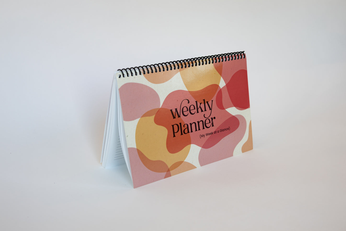 A spiral-bound weekly planner standing upright on a white surface against a white background. The cover features a retro pattern of overlapping organic shapes in pink, orange, and red, with the text 'Weekly Planner' and 'The Power of Discipline' printed on it.