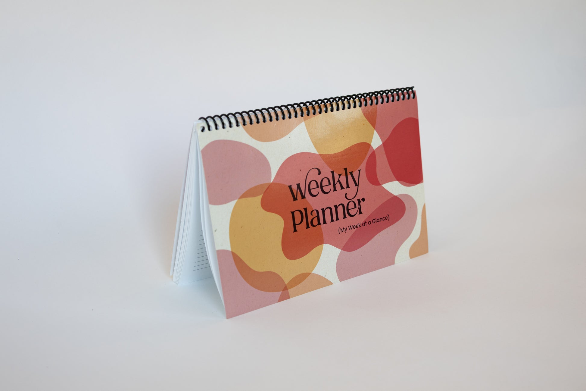 A spiral-bound weekly planner standing upright on a white surface against a white background. The cover features a retro pattern of overlapping organic shapes in pink, orange, and red, with the text 'Weekly Planner' and 'The Power of Discipline' printed on it.