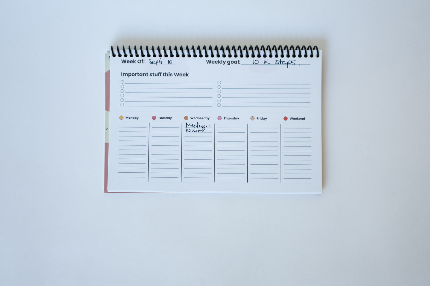 A spiral-bound weekly planner pad with lined sections for daily planning, shown open on a plain white background.
